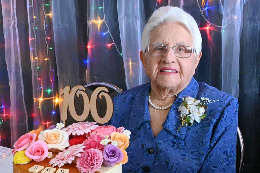 A lady smiles, sitting behind her 100th birthday cake.