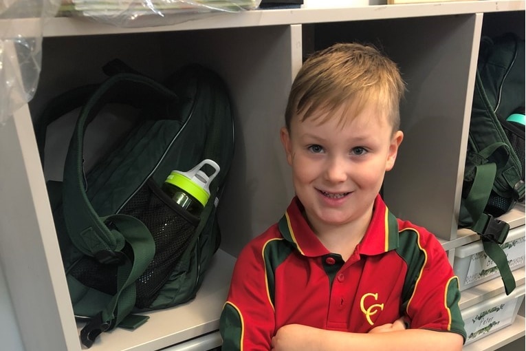 Young boy wearing school uniform standing in front of his schoolbag.