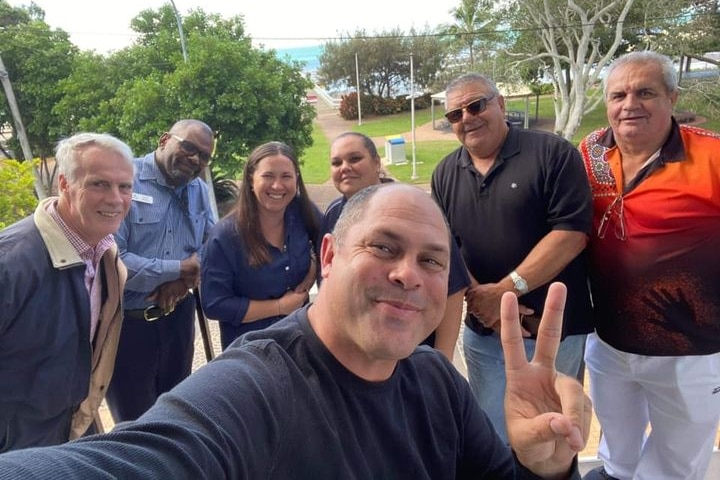 A group of men and women pose, smiling for a photo with one man in the centre making a peace sign.