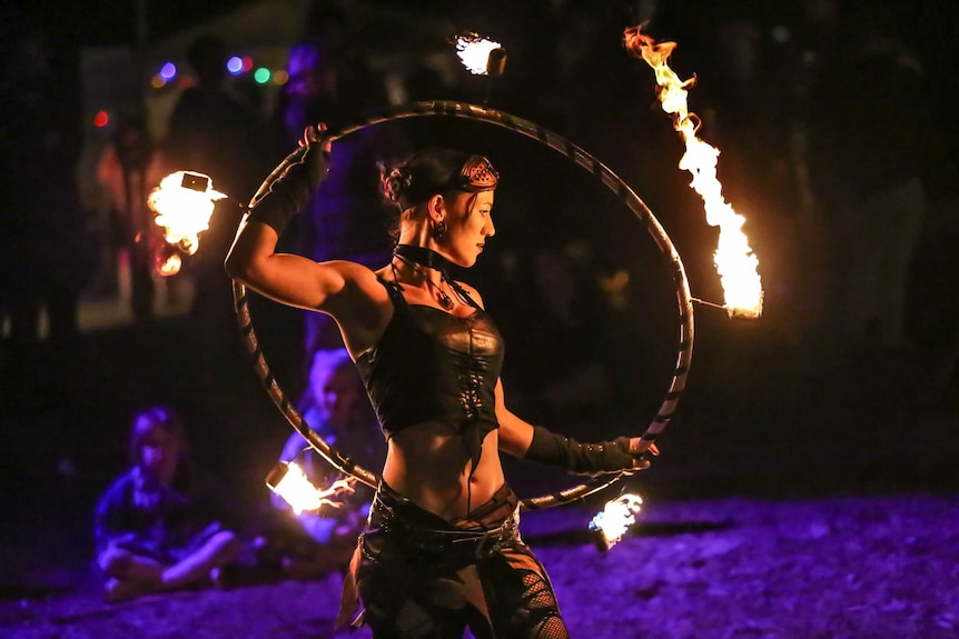 A lady on a beach at night wearing a black corset and black pants holds up a hoop that's on fire.