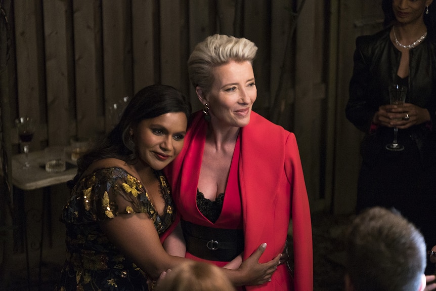 Two women, one wearing dress with embroidery, the other a red pant suit, embrace and pose for cameras and small crowd.