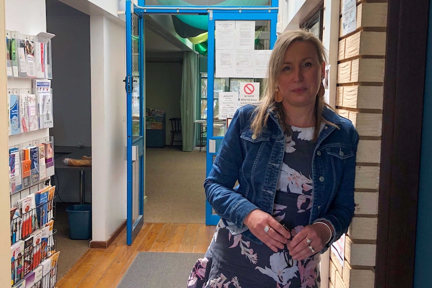 A blonde woman wearing a denim jacket and floral dress leans against a brick wall in a hallway