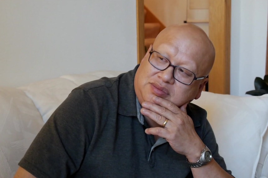 A bald man of Chinese appearance wearing glasses, a watch and a wedding ring sits thinking.