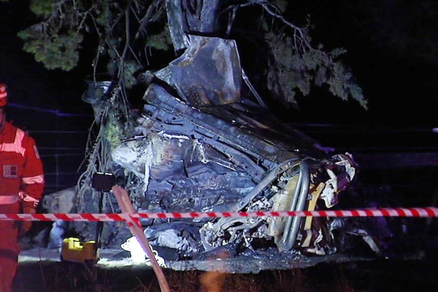 Flashlights illuminate the twisted, burnt wreckage of a car against a tree as emergency services crews walk around the scene.