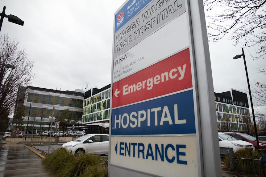 A red and blue sign saying emergency, hospital entrance, two large square buildings, car parks in the background. Overcast sky.