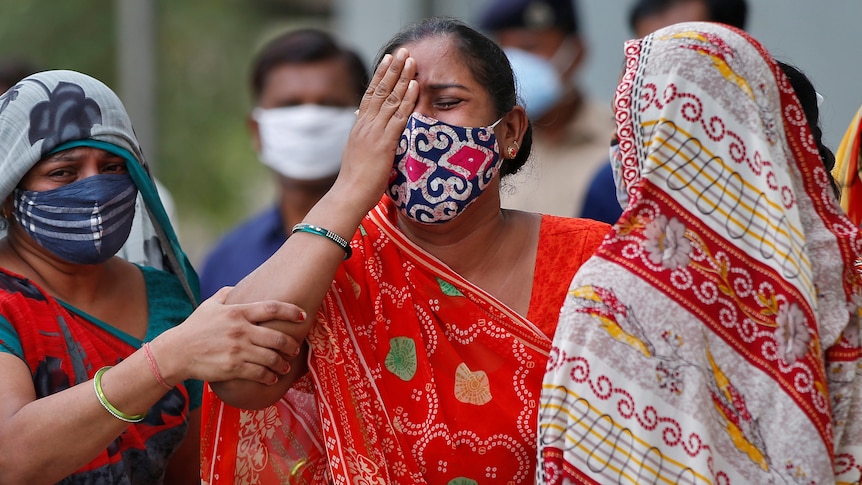 A woman in a red sari and a face mask is comforted by other women.