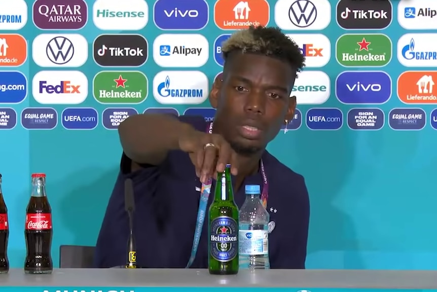 Paul Pogba reaches out to pick up a bottle of Heineken beer in front of a wall of sponsor logos.