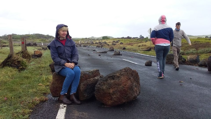 a woman sits on a large boulder which is lying across the road.