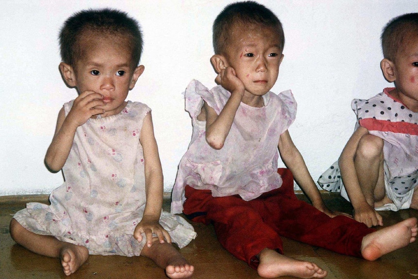 Three small, very malnourished Korean children sitting against a white wall