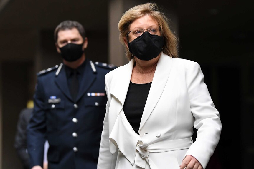 A middle-aged woman wearing a face mask and a while suit walks ahead of masked police officer.