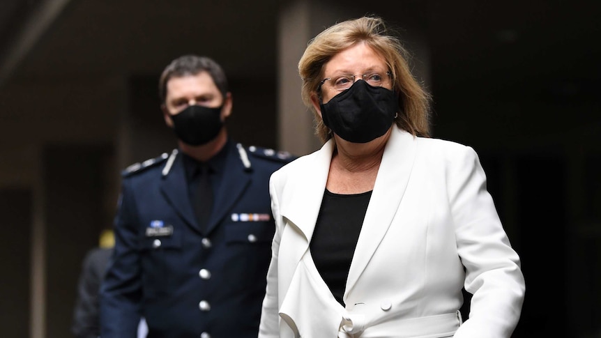 A middle-aged woman wearing a face mask and a while suit walks ahead of masked police officer.