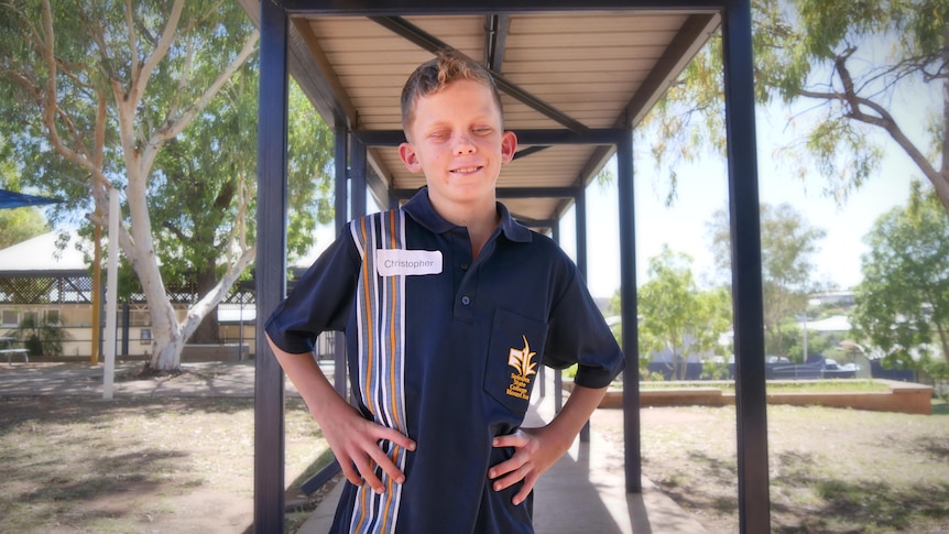 Young boy in school uniform stands with hands on hips smiling. Boy is vision impaired.