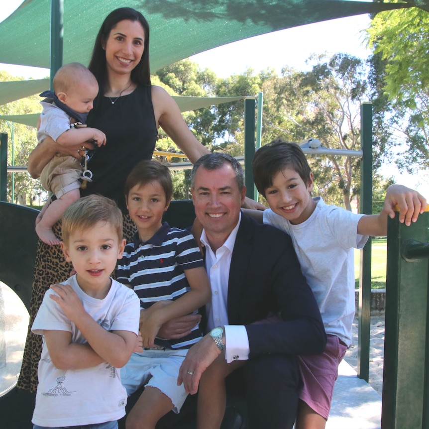 A family with a mother and father and four young sons, including a baby, smile together on a playground.