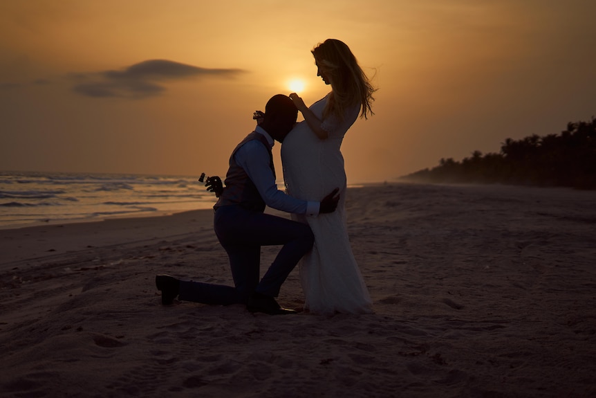 Géraldine is heavily pregnant on the beach at sunset and her husband is kissing her stomach.
