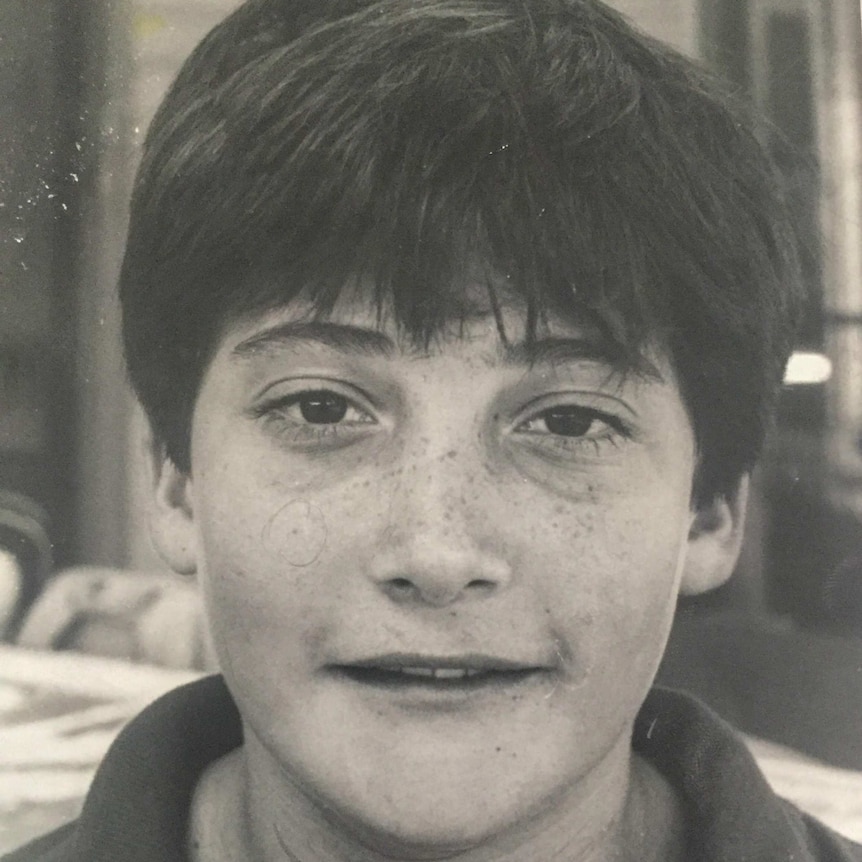 Tristan Naudi, as a young boy with freckles and short hair looks into the camera.