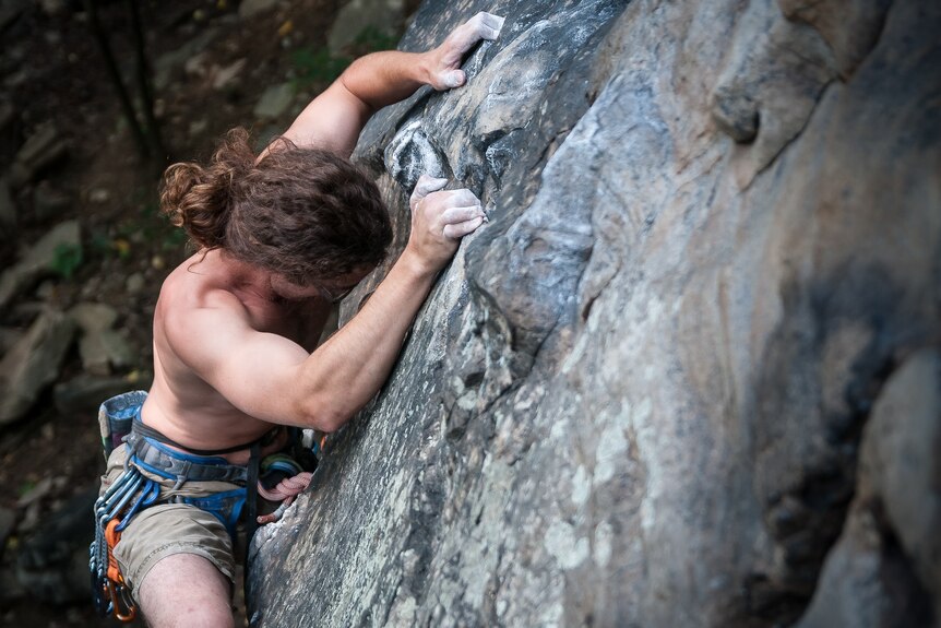 From above, a shirtless man is seen clinging to the face of a cliff, facing downwards.