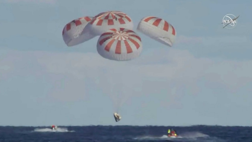 A space capsule overs above the sea with four parachutes as two small boats approach it.