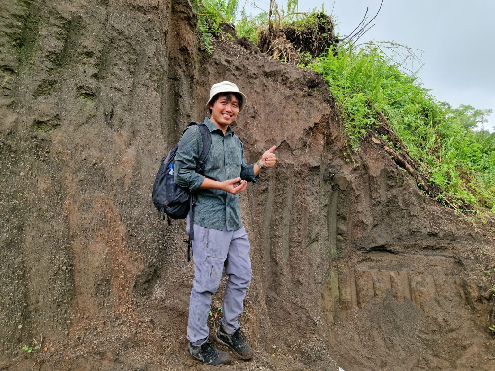 A man wearing a hat anf carrying a bag standing near an exposed earth wall