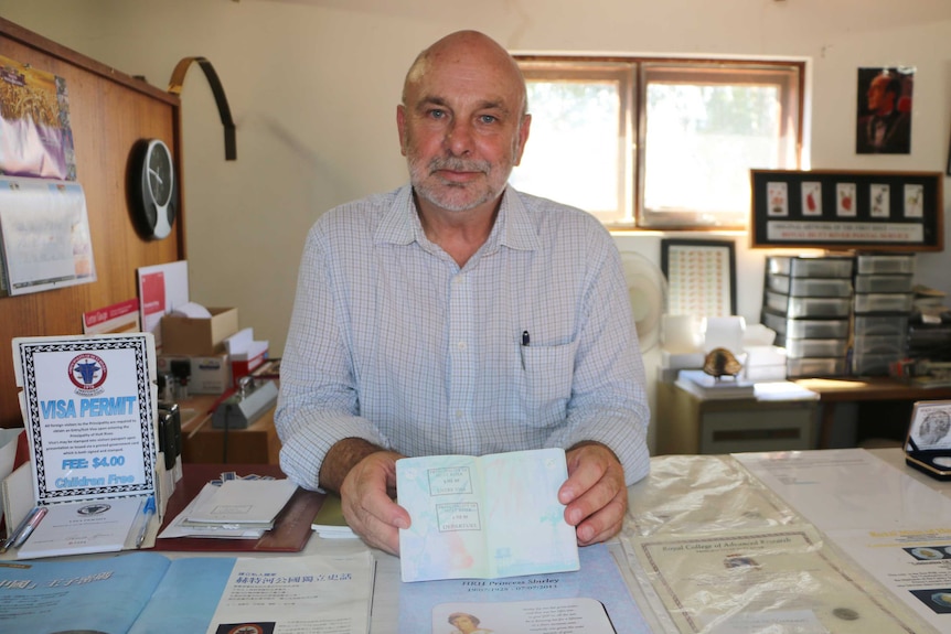 A man stands at a counter posing for a photo with a passport held out on the desk.