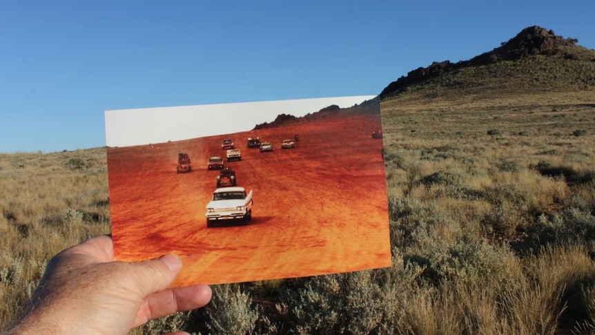 A hand holding a picture of cars on red dirt with the pinnacles in Broken Hill in the background.