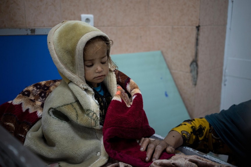 A sad looking toddler in winter clothes looks down as an adult's hand provides comfort in scruffy hospital ward