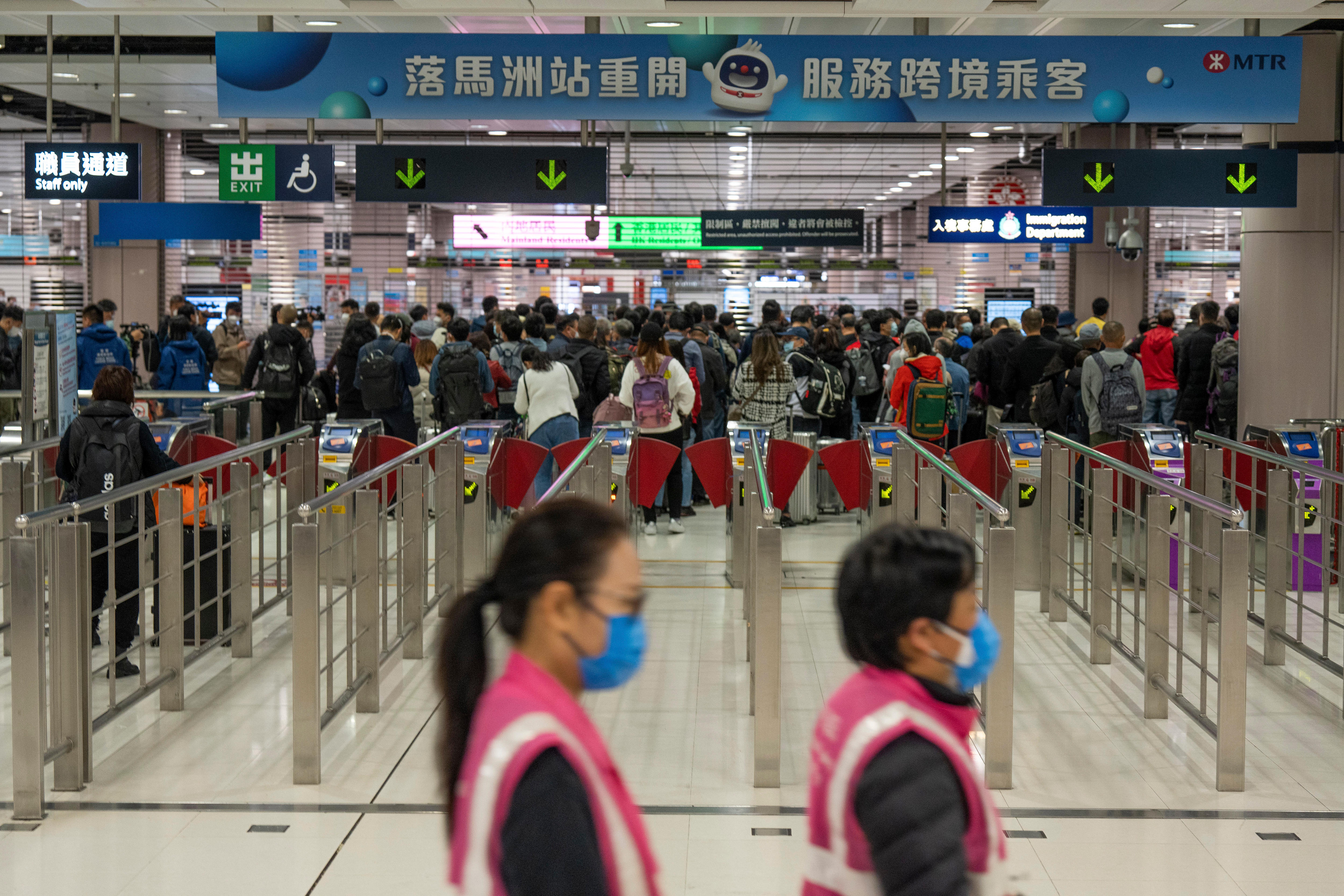 Workers wearing face masks walk by as a large group of travelers wait at a departure hall.