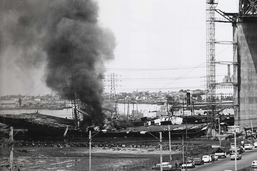 A black and white photograph of the collapsed West Gate Bridge with a fire burning in the wreckage.