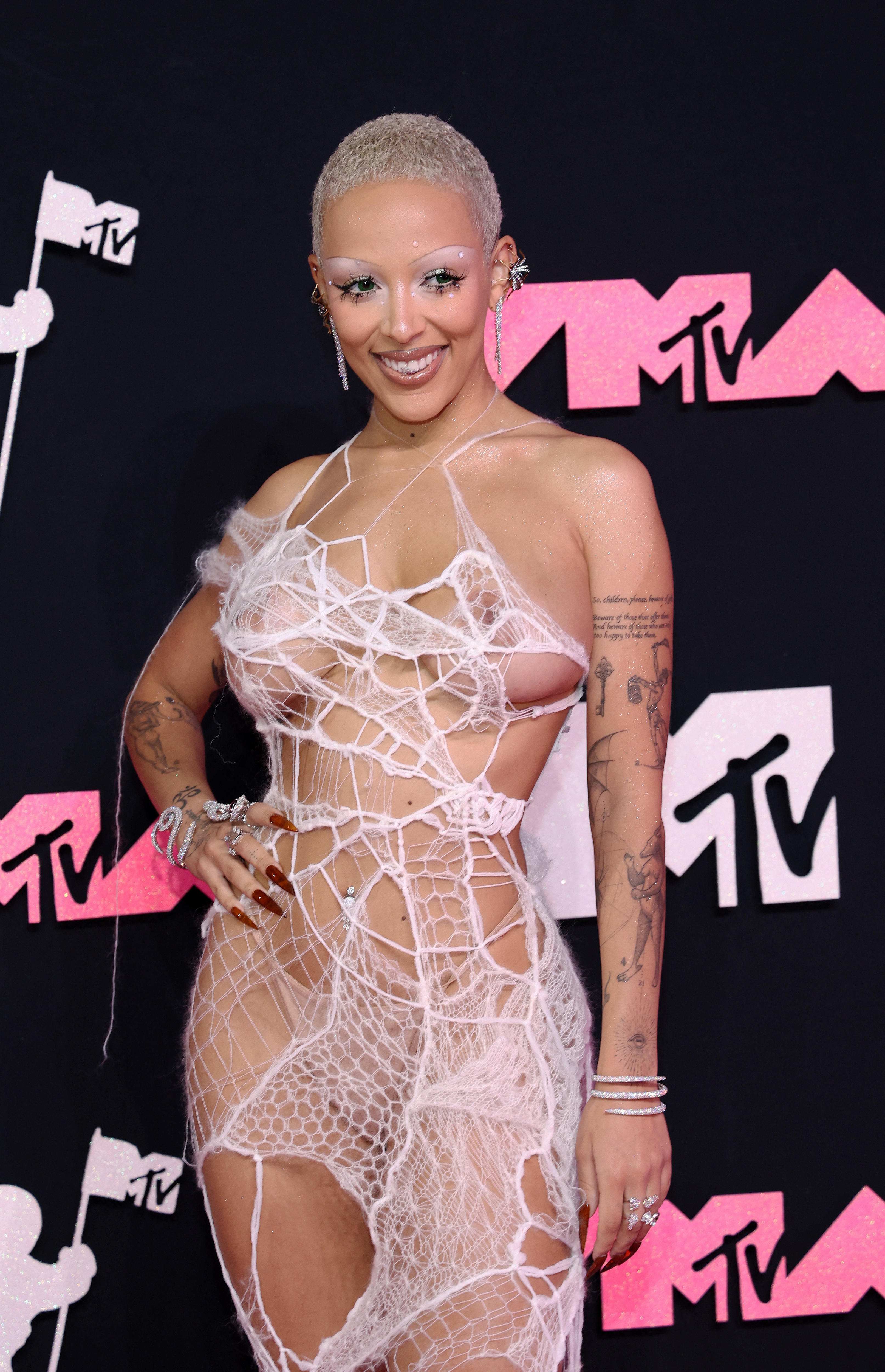 Doja Cat is pictured on a red carpet wearing a spiderweb-like dress. She smiles with one hand on her hip.