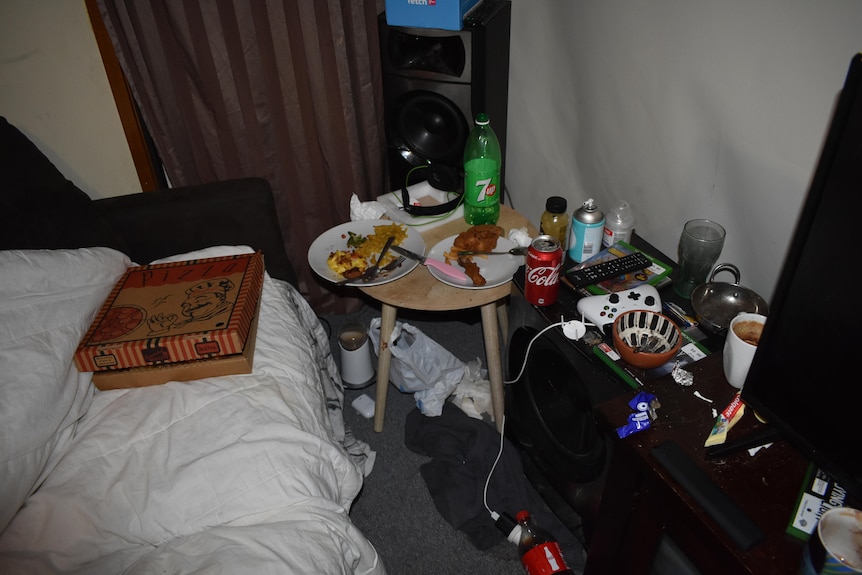 A closed pizza box on a bed next to a crowded table with drinks, and a gaming controller.