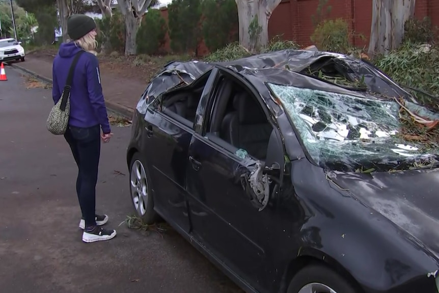 A woman looks at a black car with a crushed roof and smashed windows
