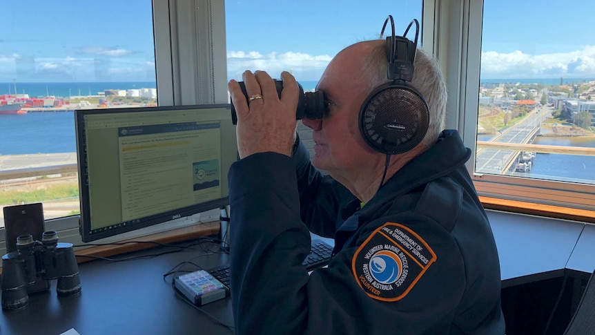 A man in a control tower looks through binoculars, with high views of Fremantle Port out the windows in the background.