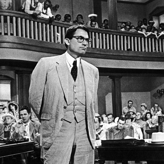 A black and white still featuring a man in a three-piece suit and thick-rimmed black glasses looking pensive in a courtroom.