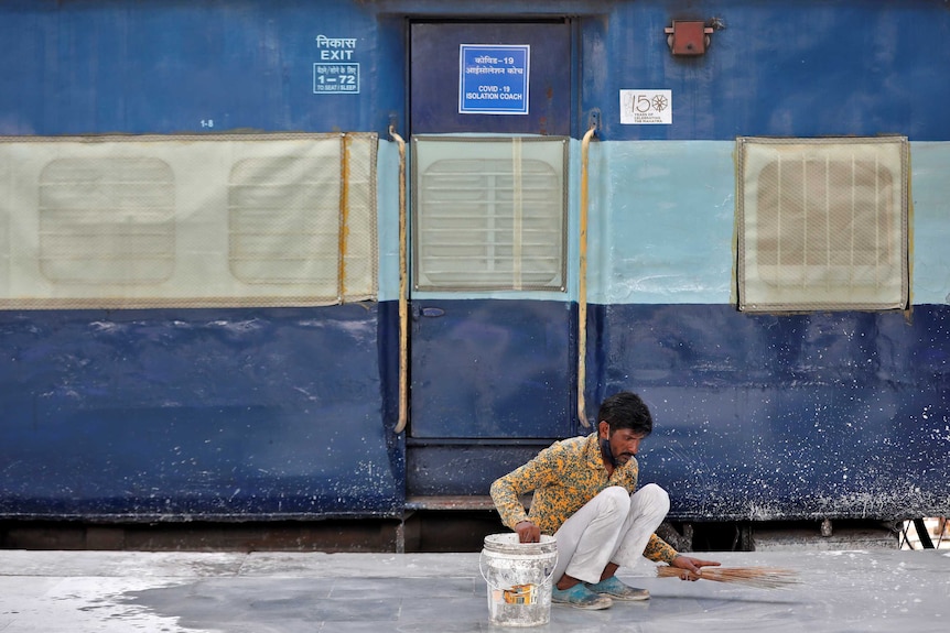 A worker crouches on the ground to clean a platform next to a net covering doors and windows of a blue parked passenger train.