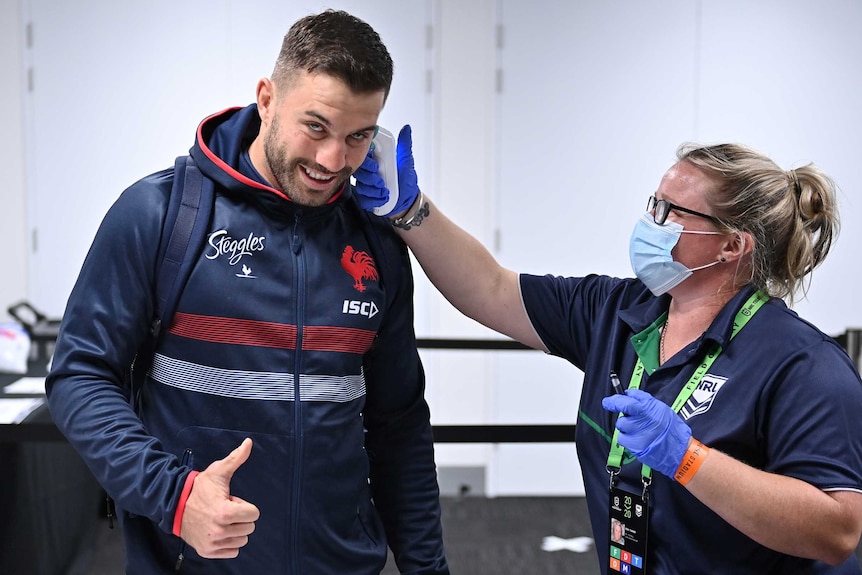 A woman puts a thermometer in the ear of Sydney Roosters fullback James Tedesco, giving a thumbs up, before an NRL game.