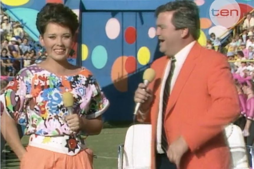 Still from 1987 video male and female host with microphones on a field and plenty of people and colour behind them
