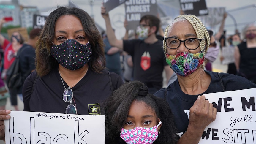 Two black adult women wearing black clothes and colourful masks stand with a small girl holding signs