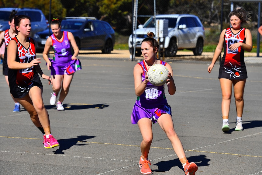 A woman wearing a purple netball uniform holding a netball, ready to pass. 