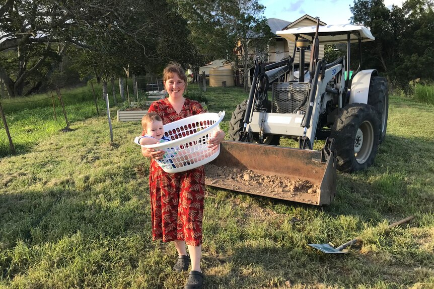 Women holding laundry basket with child in it, stands in front of a tractor