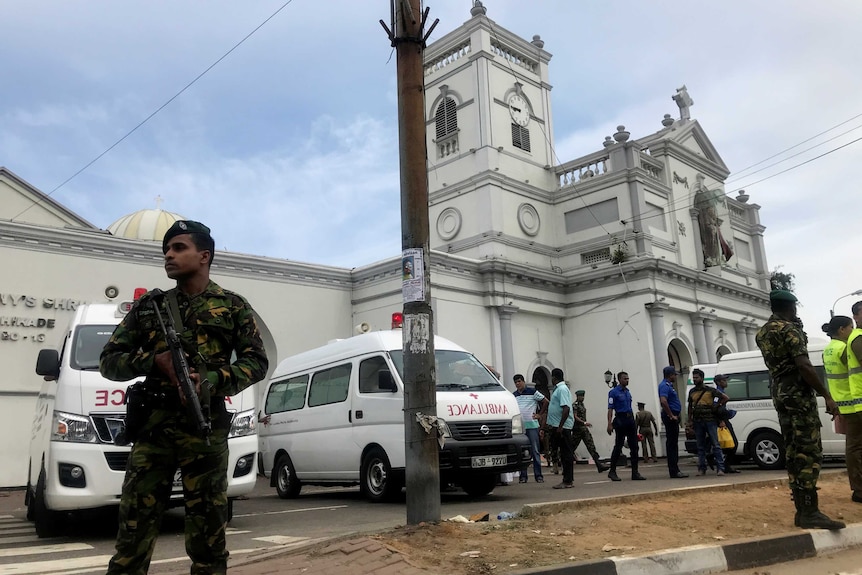 Men in military uniforms armed with guns stand in front of a church building, with ambulances stationed out the front.