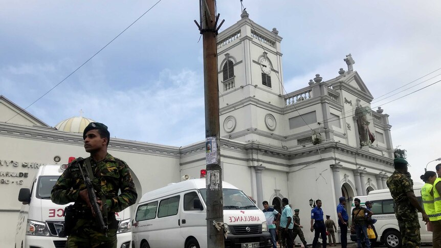 Men in military uniforms armed with guns stand in front of a church building, with ambulances stationed out the front.