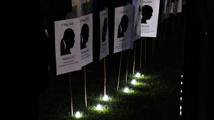 White placards with silhouettes of women are lit up by small white candles on grass.