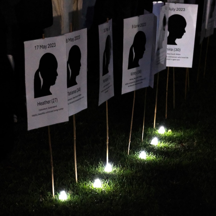 White placards with silhouettes of women are lit up by small white candles on grass.