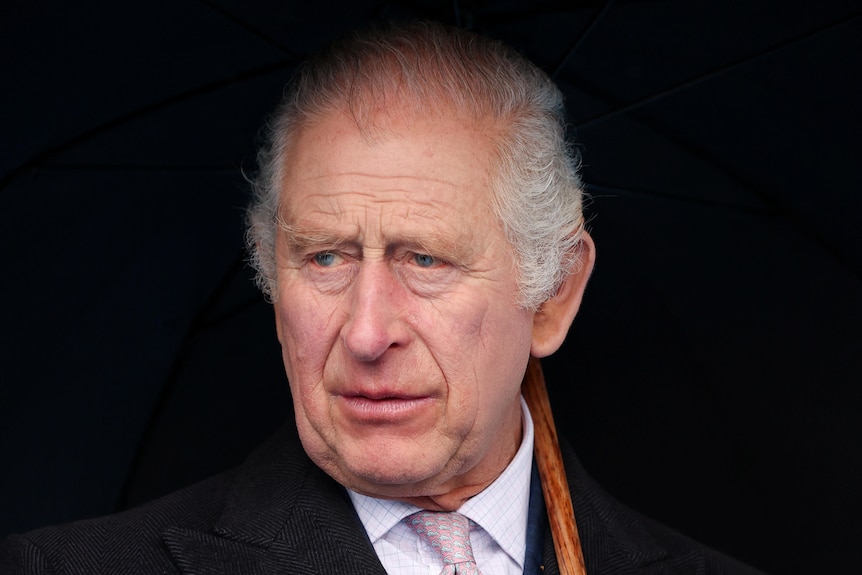 King Charles, with a stern expression on his face, looks out from under a black umbrella
