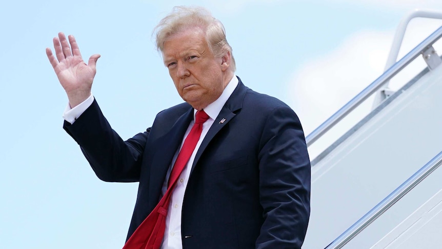 A man in a blue suit with a red tie waves his right hand as he exits an aircraft.