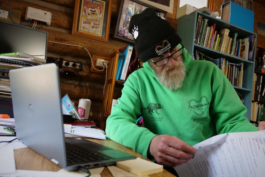 Bearded man in bright green jumper reading a page of text while sitting at his desk, bookshelves behind.