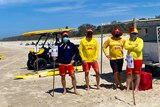 Four people wearing surf life saving uniforms stand on a beach wearing masks, surrounded by surf life saving equipment.