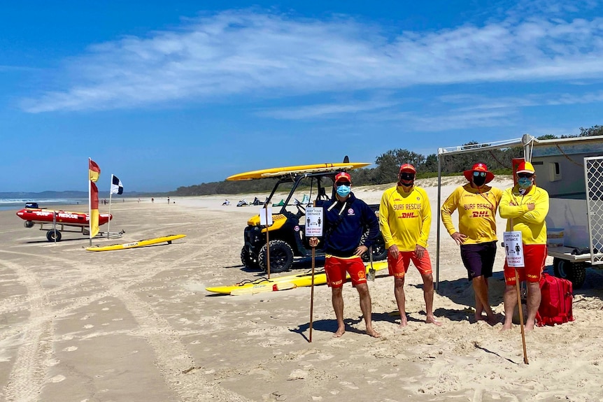 Four people wearing surf life saving uniforms stand on a beach wearing masks, surrounded by surf life saving equipment.