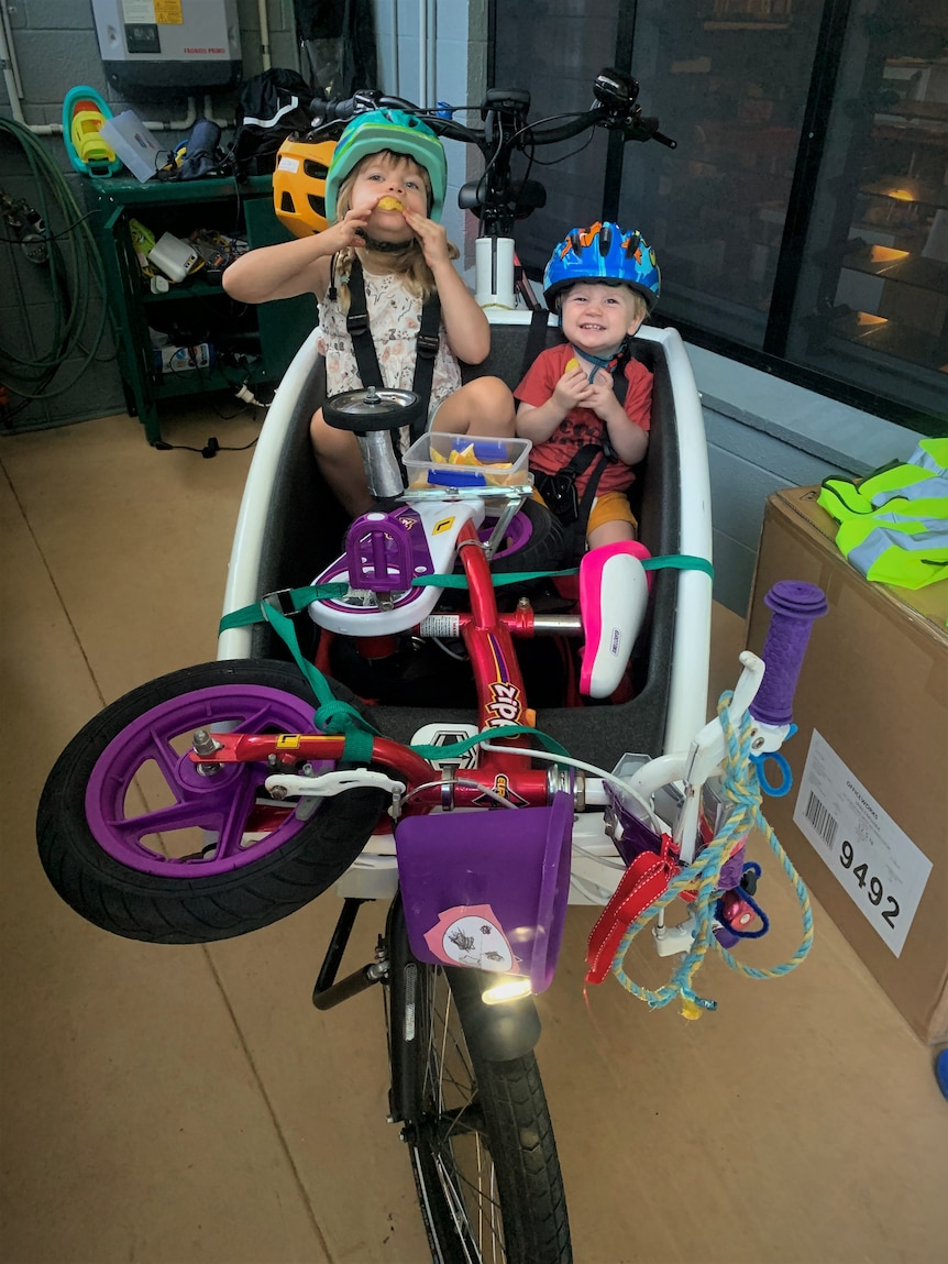 Two kids eating oranges in a cargo box bike with a purple kid's bike strapped to the front.