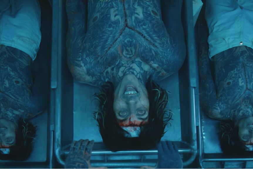 Three clones of Oli Sykes lie shirtless and pale on separate morgue trays with dead-looking eyes.