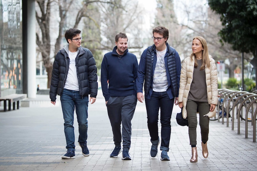 Three adult brothers in navy jackets walk along a street with their mother